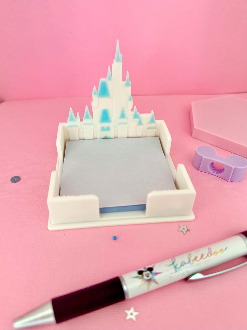 Puede incluir: Un sujetapapeles blanco en forma de castillo con detalles azules, que contiene un bloc de notas gris. Tambi&eacute;n hay un bol&iacute;grafo con la palabra "Kaleedos" y un dise&ntilde;o de Mickey Mouse. El fondo es rosa.