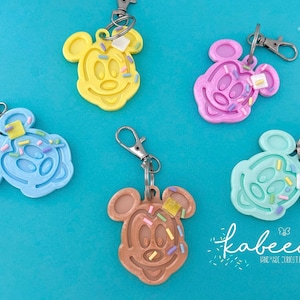 May include: Five colorful Mickey Mouse-shaped keychains with a sprinkle and frosting design. Each keychain has a different color: yellow, pink, blue, brown, and mint green. They are attached to silver-colored lobster claw clasps.