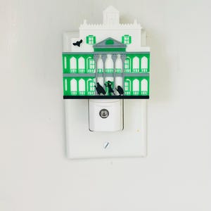 May include: A decorative night light featuring a green and white mansion design. The night light has a detailed facade with columns, windows, and a small tower. Black silhouettes of figures are visible on the front. The night light plugs into a standard wall outlet.