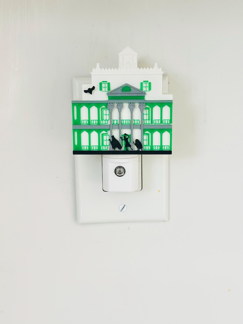 May include: A decorative night light featuring a green and white mansion design. The night light has a detailed facade with columns, windows, and a small tower. Black silhouettes of figures are visible on the front. The night light plugs into a standard wall outlet.