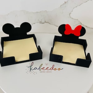 Puede incluir: Dos sujetapapeles negros, uno con orejas de Mickey Mouse y otro con orejas de Minnie Mouse y un lazo rojo. Cada soporte contiene una pila de notas adhesivas amarillas. Los soportes est&aacute;n sobre una superficie blanca.