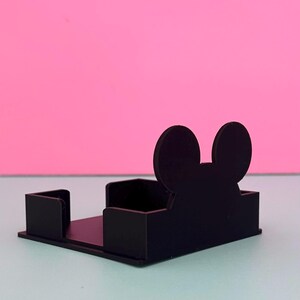 Puede incluir: Un tarjetero negro, rectangular, con un dise&ntilde;o de silueta de Mickey Mouse. El soporte tiene una base plana y dos lados verticales. El fondo es una superficie azul claro con un fondo rosa.