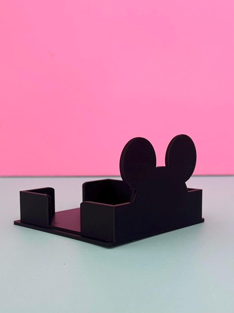 Puede incluir: Un tarjetero negro, rectangular, con un dise&ntilde;o de silueta de Mickey Mouse. El soporte tiene una base plana y dos lados verticales. El fondo es una superficie azul claro con un fondo rosa.