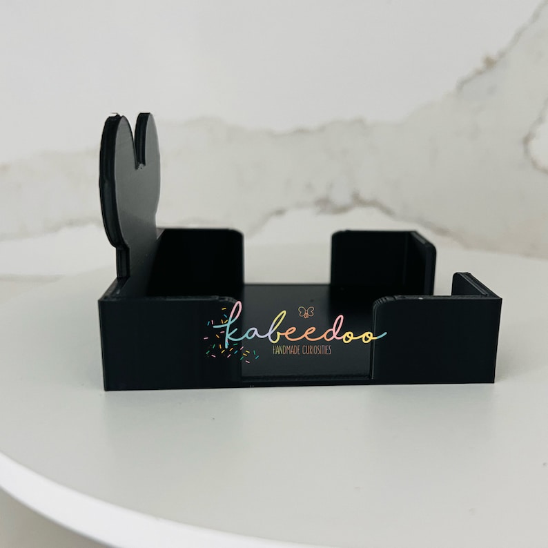 Puede incluir: Un tarjetero de acr&iacute;lico negro con un dise&ntilde;o &uacute;nico. El soporte presenta un recorte decorativo en un lado y el texto "kabeedoo HANDMADE CURIOSITIES" en una escritura colorida.