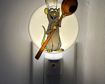 May include: A white night light with a round, translucent face featuring a cartoon chef rat holding a wooden spoon. The rat is wearing a white chef hat and has a yellow smile.