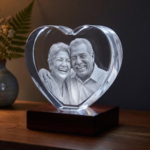 Op de afbeelding: Een hartvormig, helder glazen blok met een lasergegraveerd driedimensionaal portret van een lachend ouder echtpaar. Het glazen hart is gemonteerd op een rechthoekige, donkerbruine houten voet, wat een decoratief object vormt voor op een vlak.
