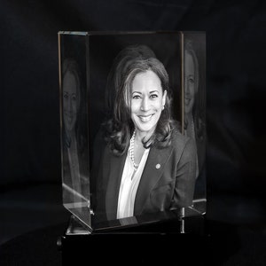 May include: A black and white 3D crystal cube with a portrait of a woman in a suit and pearl necklace. The cube is on a black base.