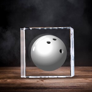 May include: A white bowling ball with three finger holes is encased in a clear, rectangular block. The bowling ball is centered within the block, which sits on a wooden surface against a dark background.