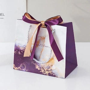 Può includere: Borsa regalo viola e bianca con un design a marmo dorato e viola. La borsa ha un manico in corda argentata e un nastro dorato con un fiocco. Il testo "Sweet Times" è stampato sulla borsa.
