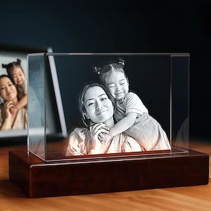May include: A 3D crystal cube with a black and white engraved image of a mother and daughter. The crystal cube is illuminated from below by a light source, creating a glowing effect. The cube is sitting on a wooden base.