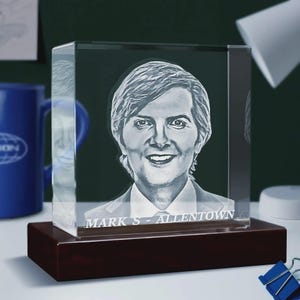 May include: A clear glass cube with a portrait of a person etched inside, mounted on a dark wooden base. The text "MARK S - ALLENTOWN" is visible at the bottom. The background includes a blue mug and office supplies.