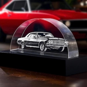 May include: A black and white etched classic car sculpture displayed under a clear, arched cover. The sculpture sits on a black rectangular base. A red car is blurred in the background.