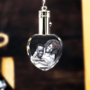 May include: A heart-shaped crystal keychain with a photo of a couple inside. The keychain is silver and has a small light on the top.