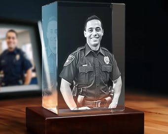May include: A rectangular glass block featuring a laser-etched 3D portrait of a smiling police officer in uniform. The clear glass block is mounted on a solid, dark brown wooden base, creating a commemorative display piece for a desk or shelf.
