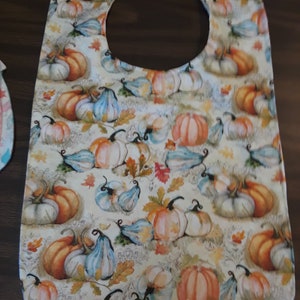 May include: A white bib with a round neck opening, featuring a pattern of orange, white, and blue pumpkins and gourds on a cream background.
