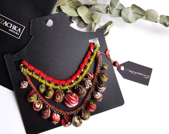 May include: A multi-strand necklace with red and green woven cords and dangling fabric-covered beads. The necklace is displayed on a black card with the brand name "Kozzachka" and a black shopping bag.
