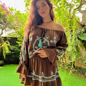 Op de afbeelding: Een bruin off-shoulder top en bijpassende rok met blauwe borduursels en kwastjes. De outfit is gemaakt van een zachte, vloeiende stof.