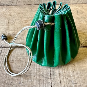 May include: A green leather drawstring pouch with a white cord and black toggle. The pouch is folded and gathered at the top, creating a rounded shape.