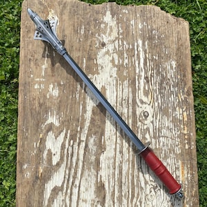 Puede incluir: Una maza de estilo medieval con una cabeza plateada con cortes decorativos y un largo mango gris oscuro. El mango está envuelto en material rojo. La maza descansa sobre una superficie de madera envejecida.