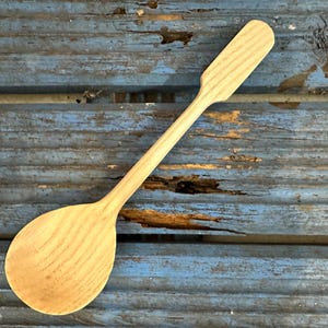 May include: A wooden spoon with a long handle and a rounded bowl. The spoon is made of light-colored wood and has visible wood grain. The handle is rectangular at the end. The spoon is likely used for cooking or serving.