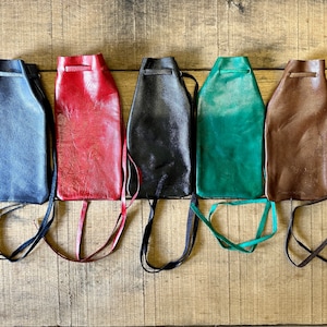 Puede incluir: Cinco bolsas de cuero en fila, cada una con cierre de cordón y correa larga. Los colores incluyen azul marino, rojo, negro, verde azulado y marrón. Las bolsas se muestran sobre una superficie de madera rústica.