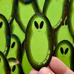 May include: A collection of oval-shaped stickers featuring a green alien design with large black eyes and a small smile. The stickers have a dark green border and the text "Wild Daisy Art".