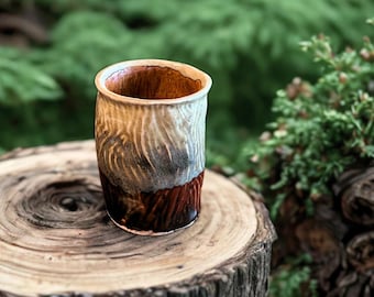 May include: A handcrafted ceramic tumbler with a textured surface. The cup features a gradient of colors, transitioning from a light beige rim to a dark brown base. The interior is a warm brown color. The tumbler is approximately 4 inches tall.