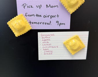 Puede incluir: Una superficie negra con dos notas y cuatro imanes amarillos con forma de raviolis. Una nota morada dice "Pick up Mom from the airport tomorrow! 9pm." Una nota blanca enumera comestibles.