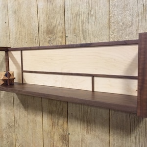 May include: A wall-mounted shelf made of walnut wood with a maple wood back panel. The shelf has a geometric design with a small wooden object on the shelf.