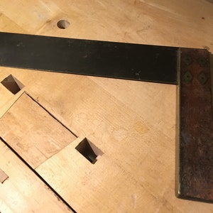 May include: A black metal try square with a wooden handle. The handle has brass accents and is worn with age.