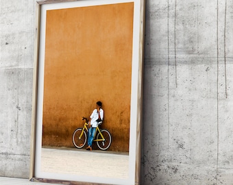 Könnte beinhalten: Gerahmter Druck mit einer Person, die sich an einem gelben Fahrrad neben einer orangefarbenen Wand anlehnt. Die Person trägt ein weißes Hemd und blaue Jeans. Das Fahrrad hat schwarze Reifen und einen schwarzen Sattel. Der Rahmen ist aus hellem Holz.