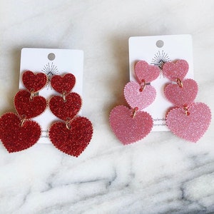 May include: Two pairs of heart-shaped dangle earrings. One pair is red glitter, the other is pink glitter. Each earring has three graduated hearts connected by gold-colored jump rings. The earrings are displayed on white cards.