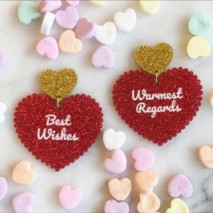 May include: A pair of red glitter heart-shaped earrings with gold glitter hearts on top. The left earring says "Best Wishes" and the right earring says "Warmest Regards".