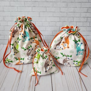 May include: Three drawstring bags with a white background and a pattern of monkeys hanging from vines. The monkeys are orange, blue, and green. The bags have orange drawstrings.