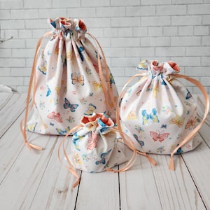 May include: Three drawstring bags in different sizes with a white background and a butterfly pattern. The bags have peach-colored drawstrings.