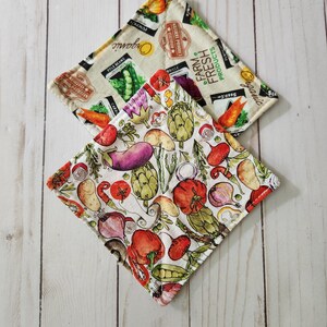 May include: Two square fabric pot holders. One is cream-colored with images of vegetables and the words "Farm Fresh Products" and "Organic". The other has a white background with colorful illustrations of various vegetables, including tomatoes, artichokes, and eggplants.