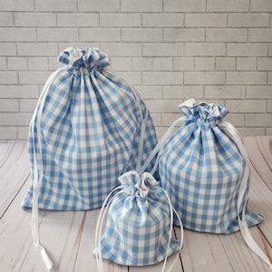 May include: Three blue and white gingham drawstring bags of varying sizes. Each bag has a gathered top and white ribbon drawstring closure. The bags are arranged on a light wood surface with a white brick background.
