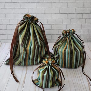 May include: Three fabric drawstring pouches in varying sizes, featuring a vertical striped pattern in shades of olive green, teal, mustard yellow, and dark brown. Each pouch is cinched at the top with a smooth, dark brown satin ribbon tie.