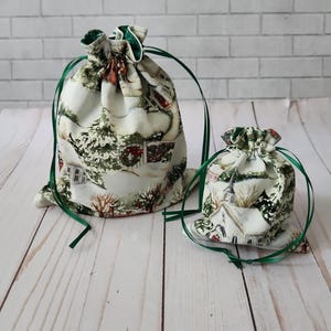 May include: Two fabric drawstring pouches in varying sizes, featuring a winter village scene with snow-covered houses, evergreen trees, and wreaths. The bags are made of off-white fabric with green ribbon closures and a green interior lining.