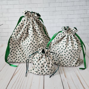 May include: Three green and white drawstring bags with a Christmas tree pattern. The bags are different sizes and are arranged in a row on a white wooden surface.