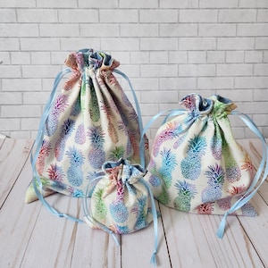 May include: Three drawstring bags made of fabric with a pineapple print in shades of blue, green, yellow, orange, and pink. The bags are lined with a light blue fabric and have light blue drawstrings.