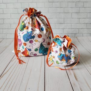 May include: Two drawstring bags with orange ribbon ties. The bags are made of white fabric with a colorful print of cats playing with yarn balls. The larger bag is in the background and the smaller bag is in the foreground.