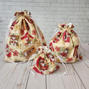 May include: Three fabric gift bags with a snowman and star pattern. The bags are in different sizes and are made of a cream-colored fabric with red, green, and yellow accents.
