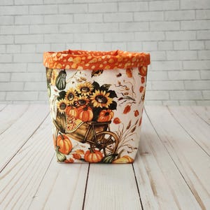 May include: A fabric storage basket with a fall harvest theme. The basket features a print of sunflowers, pumpkins, and a wheelbarrow on a white background. The top edge is trimmed with an orange and white polka dot fabric.