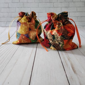 May include: Two small fabric drawstring bags with autumnal leaf patterns. One bag is yellow, orange, and brown with a leaf pattern. The other bag is red, orange, and green with a floral pattern.