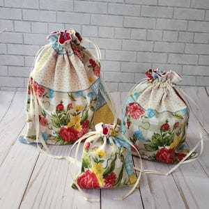 May include: Three drawstring pouches of varying sizes made from fabric featuring a floral pattern with red, yellow, and green flowers on a cream background, accented with light blue and yellow polka dot panels, each closed with thin white ribbons.