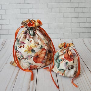 May include: Two drawstring bags with a floral pattern featuring pumpkins, sunflowers, and leaves in shades of orange, yellow, and green. The bags have orange ribbon drawstrings and are set against a white brick wall and a wooden surface.