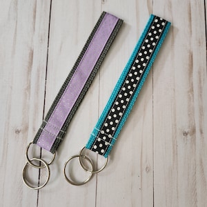 May include: Two keychains with silver key rings. One keychain has a teal and black ribbon with white polka dots. The other keychain has a gray ribbon with a purple and silver glitter stripe.