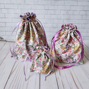May include: Three drawstring bags in different sizes with a floral print and purple ribbon ties. The bags are made of fabric and have a white background with pink, yellow, and purple flowers.