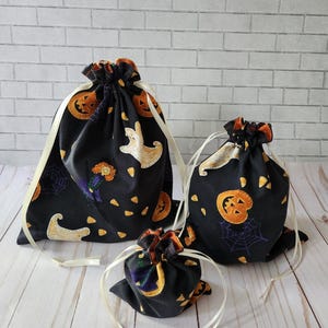 May include: Three black drawstring bags of varying sizes featuring a Halloween-themed print. The fabric pattern includes orange jack-o'-lanterns, white ghosts, purple spiderwebs, and yellow candy corn. Each bag has a cream-colored ribbon closure.